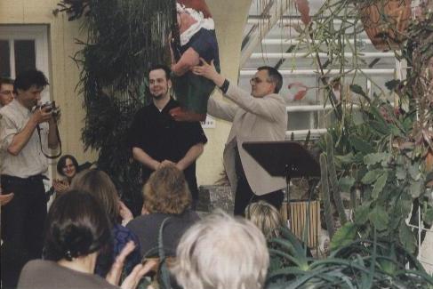 Buchautor Manfred Fock hebt eine große Gartenzwerg-Figur auf einer Bühne im Gewächshaus vor Publikum hoch.