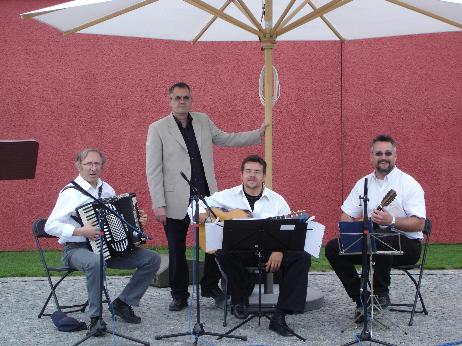 Buchautor Manfred Fock steht im hellen Sakko hinter drei sitzenden Musikern mit Akkordeon, Gitarre und Mandoline unter einem Sonnenschirm.