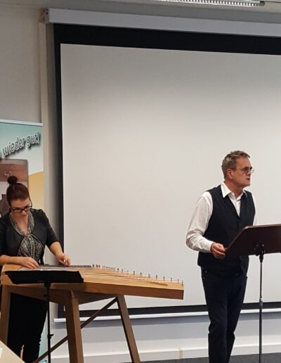 Buchautor Manfred Fock im Westen-Outfit spricht an einem Notenständer, während eine Musikerin ihn an der Zither begleitet.