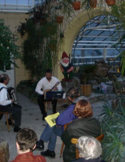 Ein Akkordeonspieler und ein Gitarrist spielen Musik bei einer Lesung in einem Gewächshaus vor sitzenden Zuhörern.