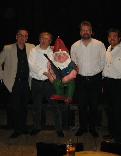 Buchautor Manfred Fock im hellen Sakko gemeinsam mit Musikern und einer großen Gartenzwerg-Figur auf der Bühne.