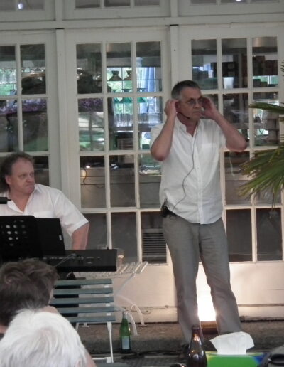 Buchautor Manfred Fock im weißen Hemd richtet sein Kopfbügelmikrofon bei einer Lesung neben einem Keyboarder.
