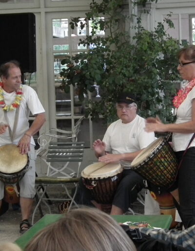 Eine Musikgruppe mit Djembe-Trommeln und Blumenketten umrahmt eine Veranstaltung von Buchautor Manfred Fock.