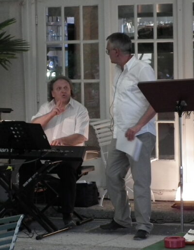 Buchautor Manfred Fock im weißen Hemd im Gespräch mit einem Keyboarder bei einer Live-Lesung.