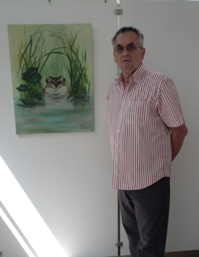 Buchautor Manfred Fock im rot-weiß gestreiften Hemd steht neben einem Gemälde, das einen Frosch im Teich zeigt.