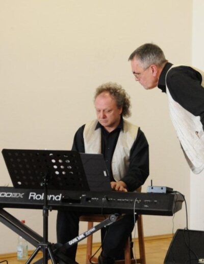 Buchautor Manfred Fock im hellen Westen-Outfit beugt sich zu einem Keyboarder am Roland-E-Piano herunter.