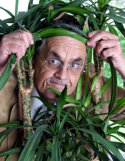 Buchautor Manfred Fock blickt mit Brille humorvoll und versteckt durch die grünen Blätter einer Yucca-Palme.