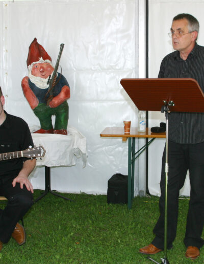 Buchautor Manfred Fock spricht bei einer Lesung in einem Festzelt an einem Holzpult, während ein Musiker ihn auf der Gitarre begleitet.