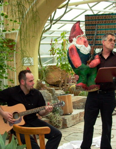 Buchautor Manfred Fock hält eine große Gartenzwerg-Figur im Arm und gestikuliert bei einer musikalisch begleiteten Lesung in einem Gewächshaus.