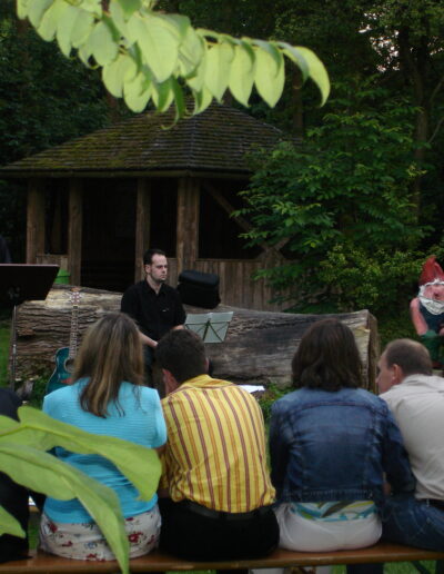 Buchautor Manfred Fock bei einer Open-Air-Lesung im Grünen mit einem Gitarristen vor sitzendem Publikum und einer Gartenzwerg-Figur.