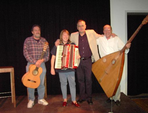 Buchautor Manfred Fock im hellen Sakko lächelt Arm in Arm mit Musikern auf einer Bühne.