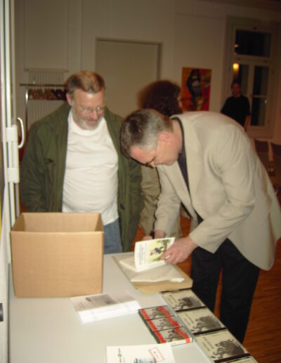 Buchautor Manfred Fock im hellen Sakko signiert ein Buch an einem Tisch für einen interessierten Leser mit Brille und grünem Parka.