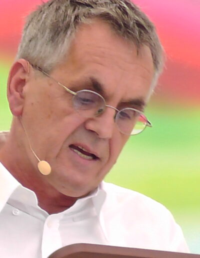 Porträt von Buchautor Manfred Fock im weißen Hemd mit Brille und Kopfbügelmikrofon bei einer Lesung.