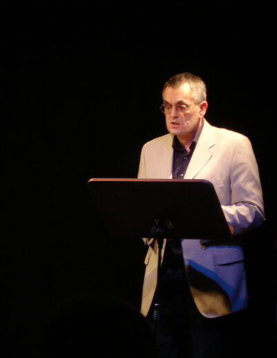 Buchautor Manfred Fock im hellen Sakko bei einer literarischen Lesung am Rednerpult vor dunklem Hintergrund.