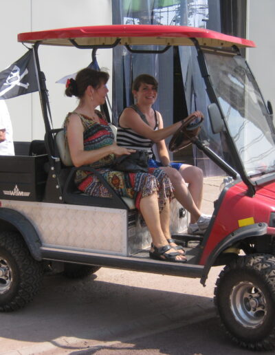 Buchautor Manfred Fock sitzt auf der Ladefläche eines roten Golfkarts mit Piratenflagge, während zwei Frauen vorne sitzen.