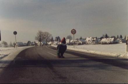 Eine Person mit einer auffälligen roten Mütze hockt mitten auf einer asphaltierten Straße in einer verschneiten Winterlandschaft.
